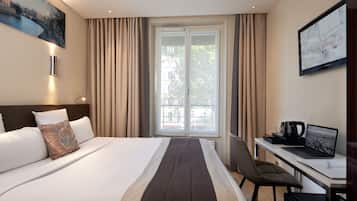 Une chambre d’hôtel avec un lit, un bureau avec un ordinateur portable, une machine à café et une grande fenêtre avec des rideaux.