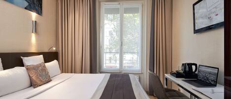 A hotel room with a bed, a desk with a laptop, a coffee maker, and a large window with curtains.