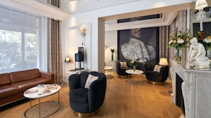 A spacious living room with a brown leather sofa, a black armchair, and a round coffee table. There are two black armchairs with lamps, a white fireplace with a statue, and a large framed artwork on the wall. - Hôtel Tourisme Avenue (Paris)