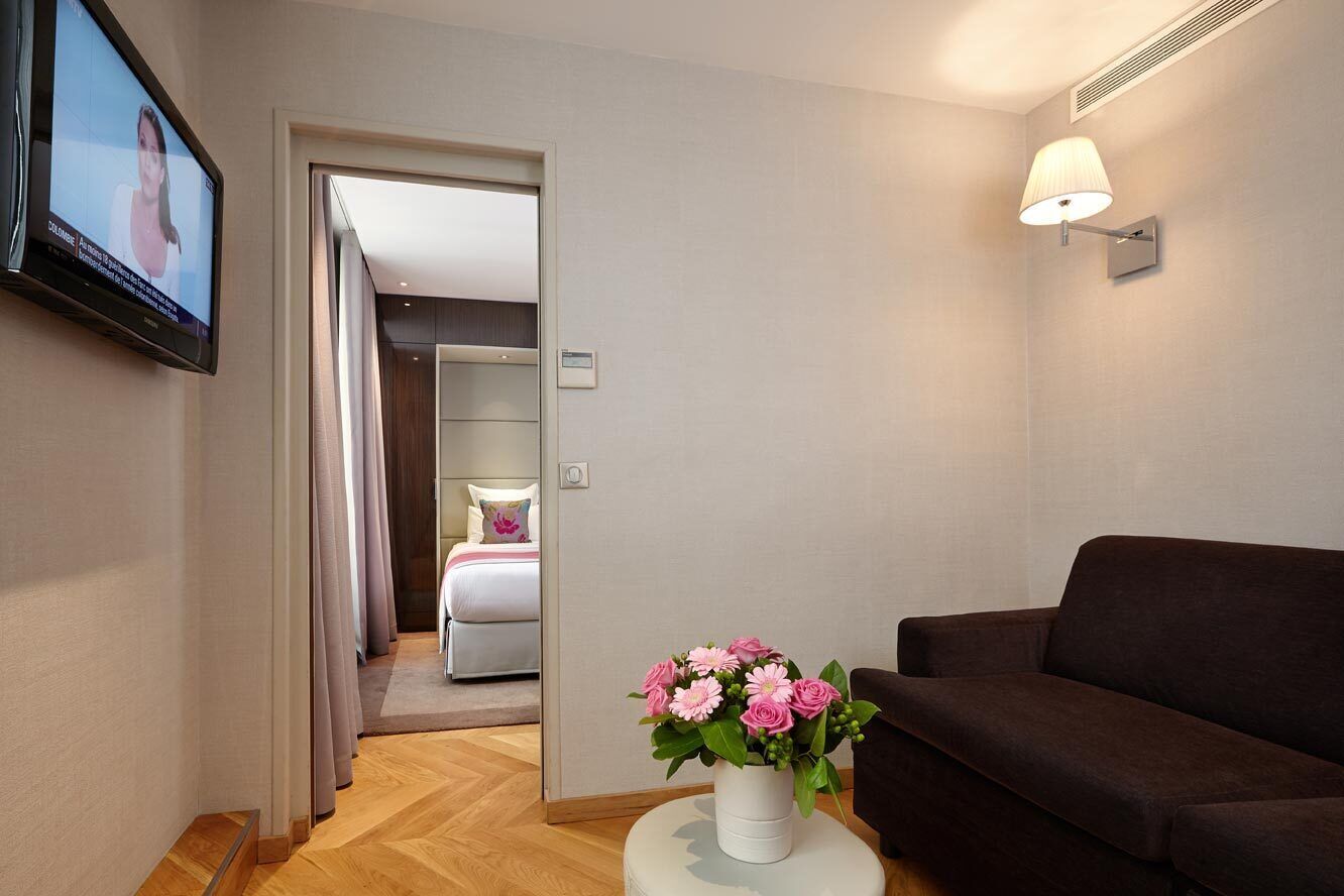 A hotel room with a dark brown sofa, a white vase with pink flowers, a television on the wall, and a bed with a white duvet and pink pillow.