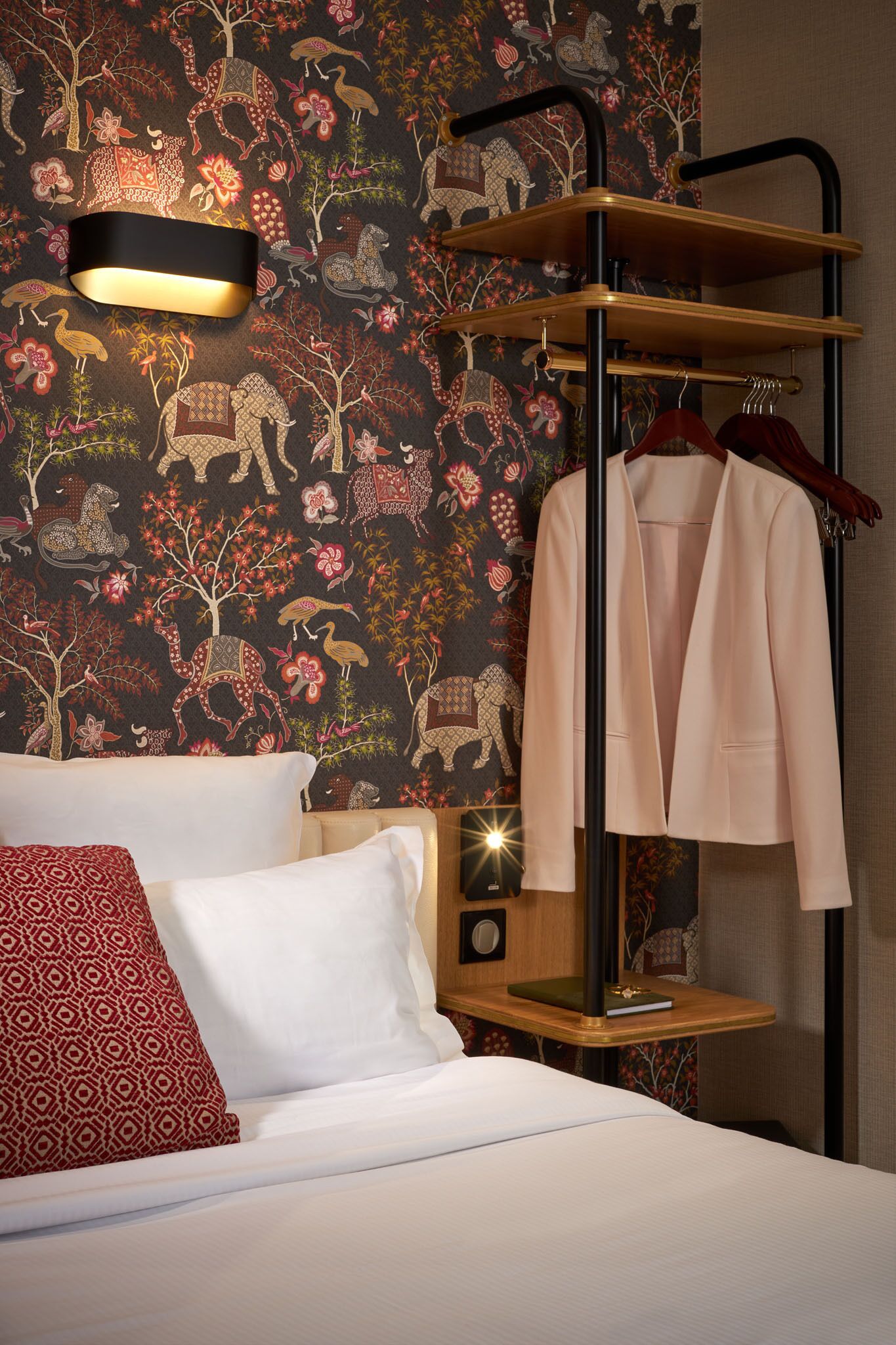 A hotel room with a bed, a wooden nightstand, a wall-mounted shelf with a coat, and a decorative wall with animal patterns.