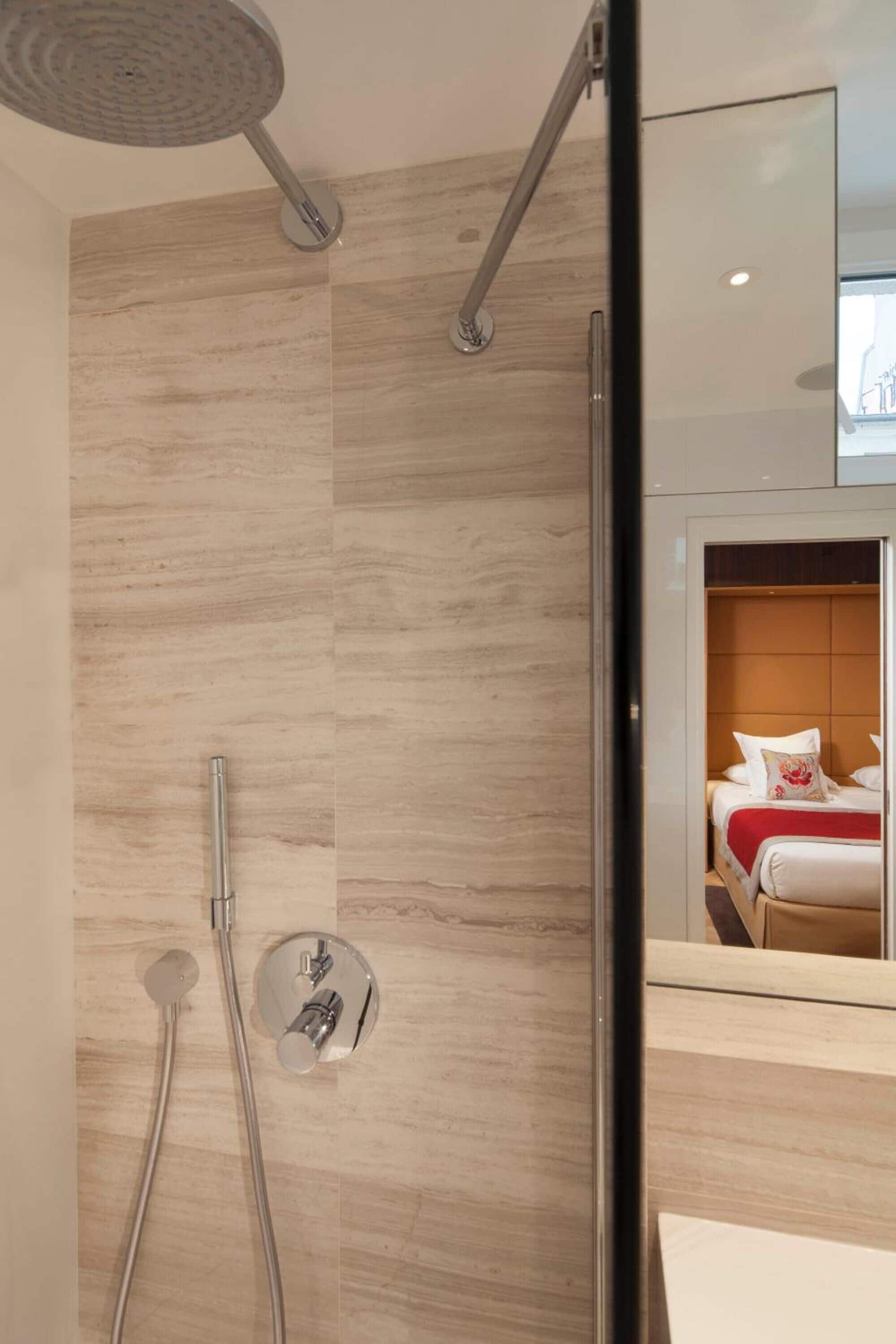 Um banheiro moderno com chuveiro, espelho e uma cama visível no reflexo.