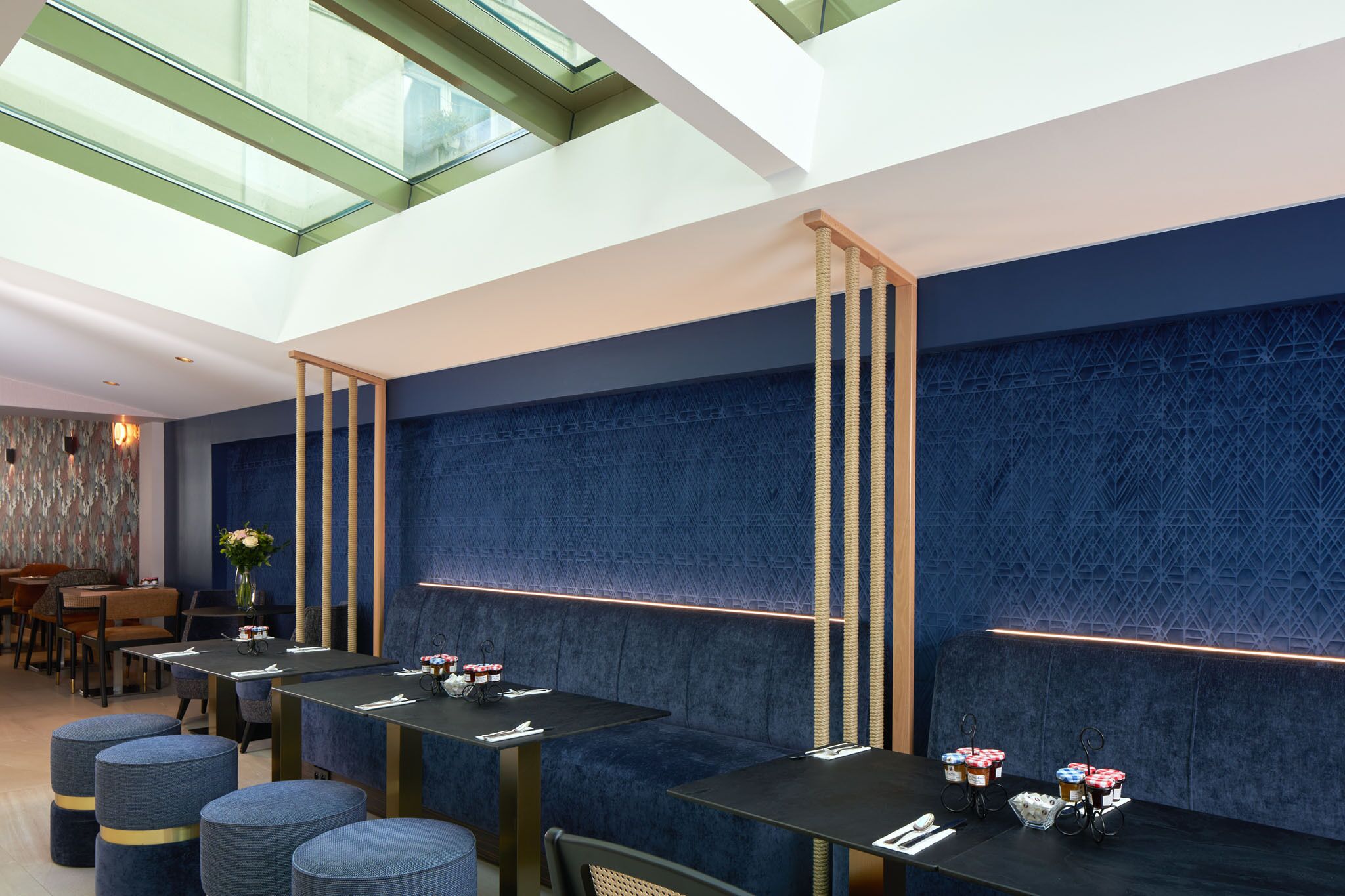 A modern restaurant with high ceilings, a skylight, and a blue textured wall. Tables are set with menus and small dessert stations.