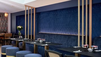 Ein modernes Restaurant mit dunkelblauen Wänden, vertikalen Holzelementen und blau gepolsterten Sitzgelegenheiten. Die Tische sind mit weißen Servietten und kleinen Obstkörben gedeckt.