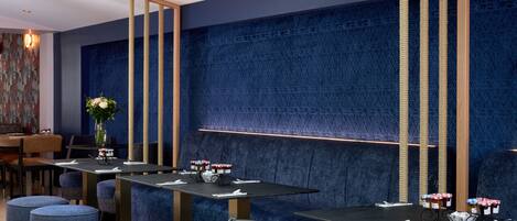 Un restaurante moderno con paredes azul oscuro, elementos verticales de madera y asientos tapizados en azul. Las mesas están dispuestas con servilletas blancas y pequeñas cestas de fruta.