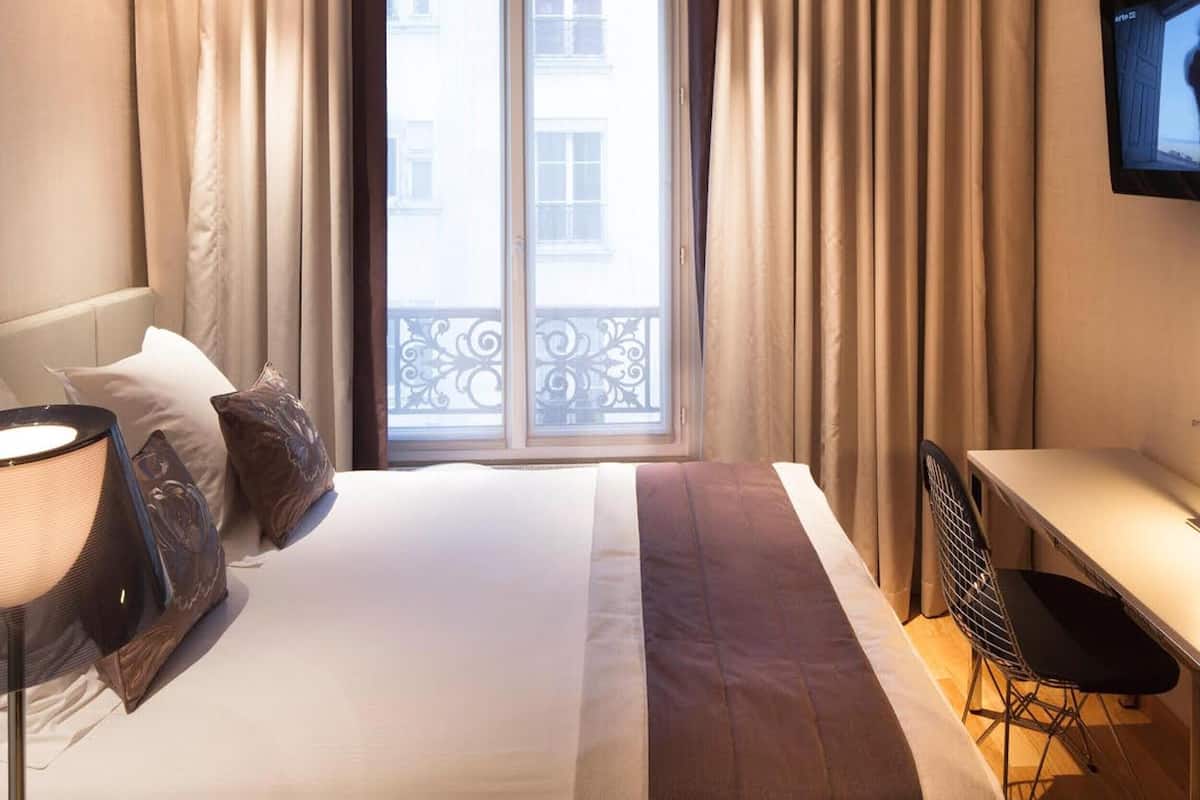 A hotel room with a bed, a desk, a chair, a TV, and a window with curtains.
