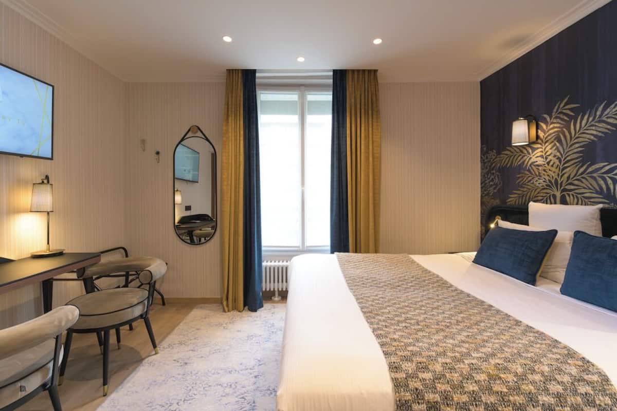 A hotel room with a bed, a desk with a chair, a large mirror, and a window with curtains.