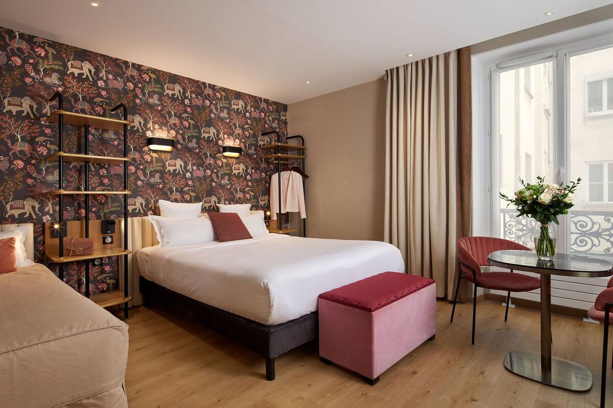 A hotel room with a large bed, a small table with chairs, a red ottoman, a decorative wall with animal patterns, and a window with curtains.