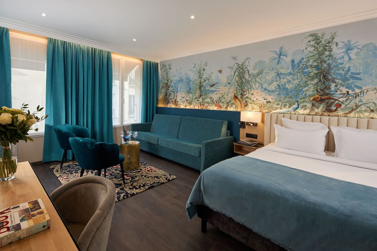 A hotel room with a large bed, a blue sofa, a small table with a vase, and a mural of a tropical scene on the wall.