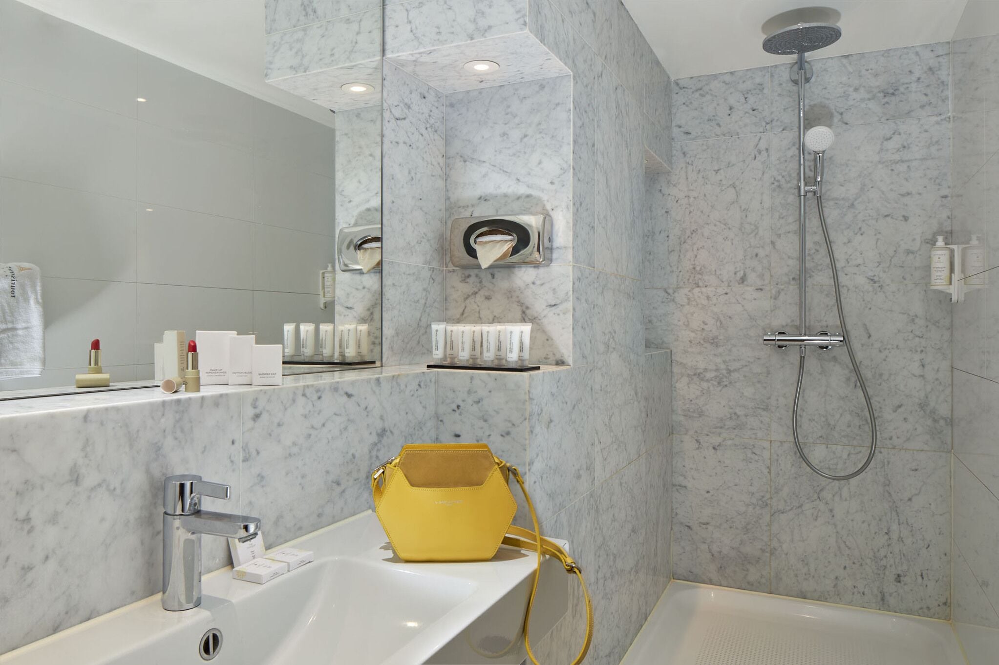 A modern bathroom with a marble wall, a sink, a mirror, a shower, and a yellow handbag on the counter.