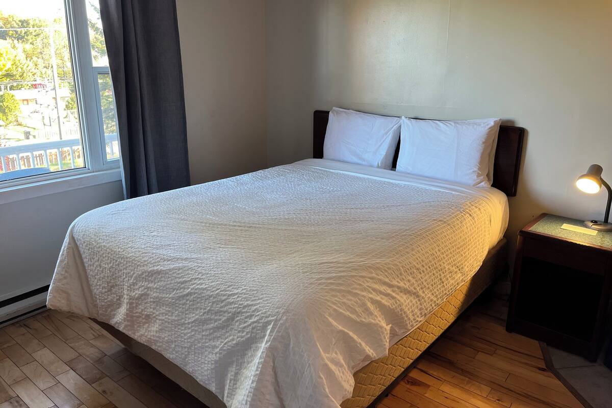 Un lit bien fait, avec du linge de lit blanc, un plancher en bois, une table de chevet avec une lampe et une fenêtre avec des rideaux.