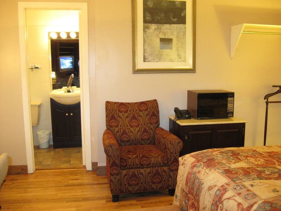 A hotel room with a wooden floor, a patterned armchair, a bed with a floral bedspread, a desk with a microwave, and a painting on the wall.