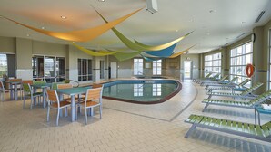 Piscina interior con sillones de descanso y mesas, rodeada de amplias ventanas y suelo de baldosas.