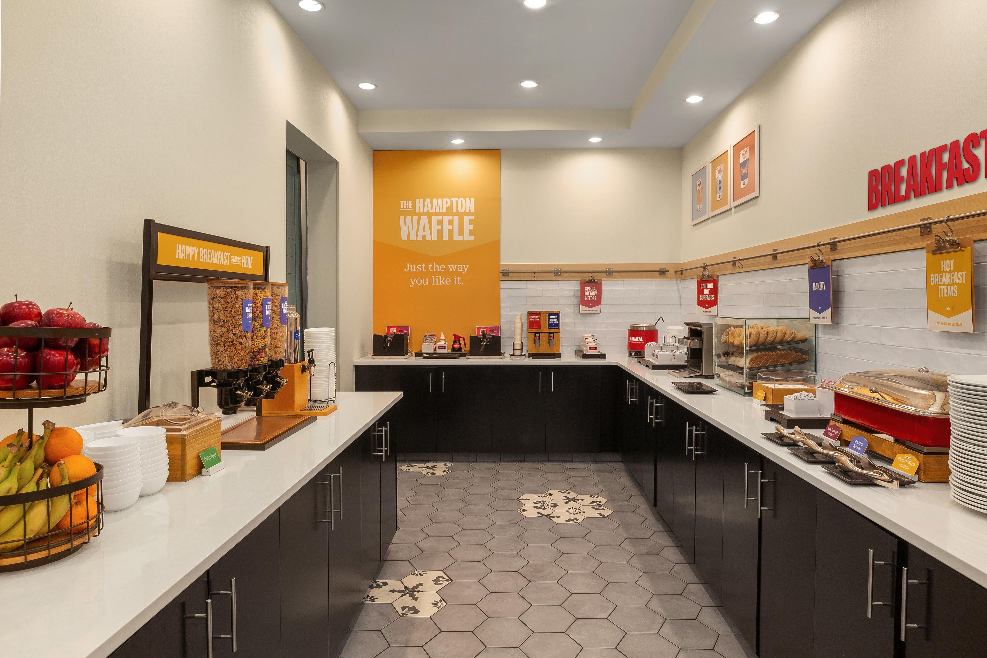 A breakfast area with a sign for The Hampton Waffle, a fruit display, and various breakfast items.