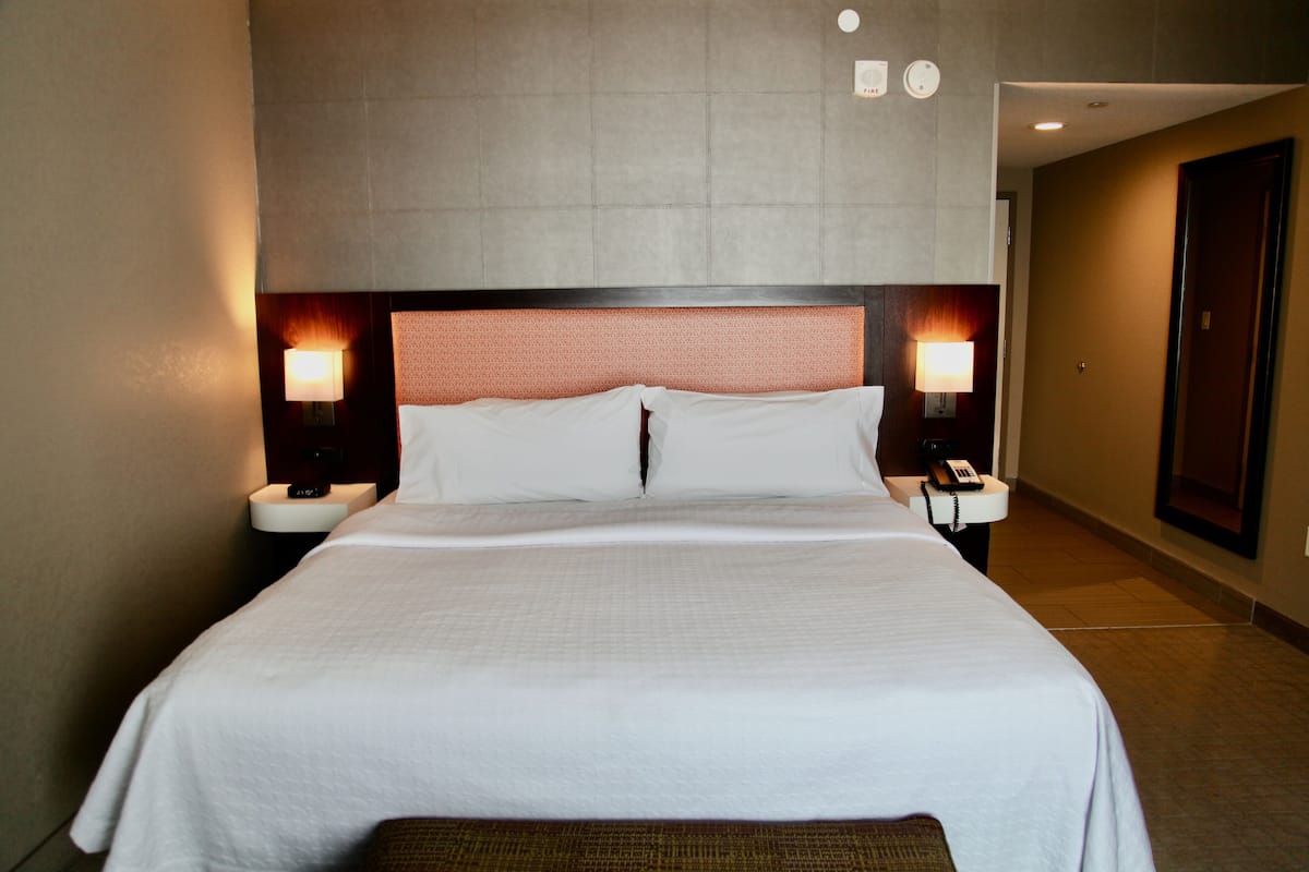 Habitación de hotel con una cama grande, dos lámparas de noche, una mesita de noche con un teléfono y una puerta a la derecha.