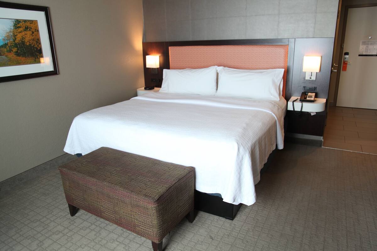 Una habitación de hotel con una cama grande, una mesita de noche con una lámpara, un cuadro en la pared y un pequeño banco.