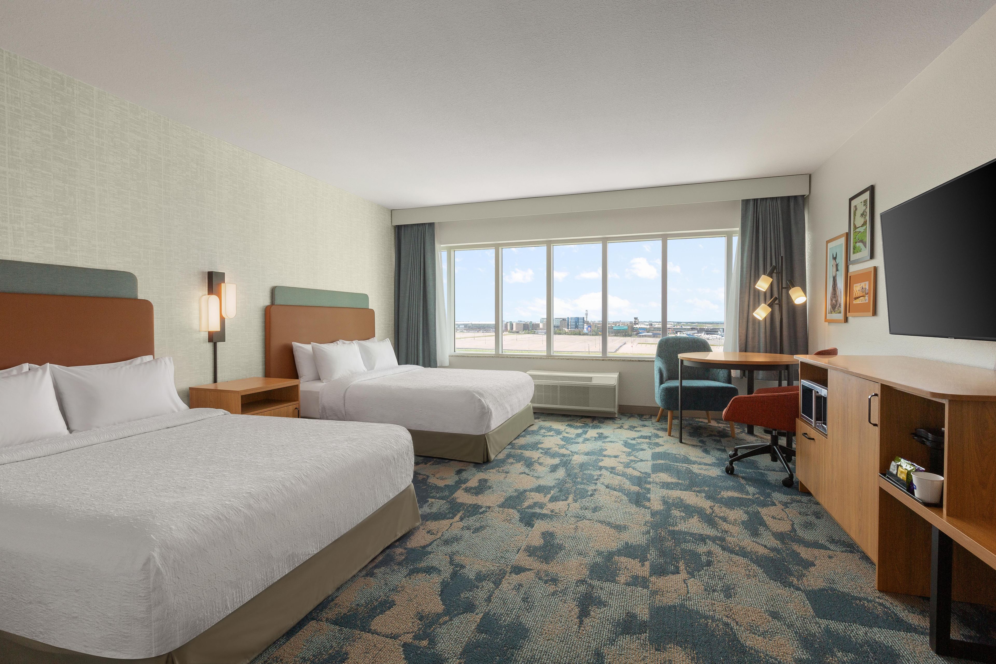 A hotel room with two beds, a desk, a chair, a TV, and a large window with a city view.