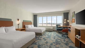 Quarto de hotel com duas camas, uma escrivaninha, uma cadeira, televisão e uma janela grande com vista para a cidade.