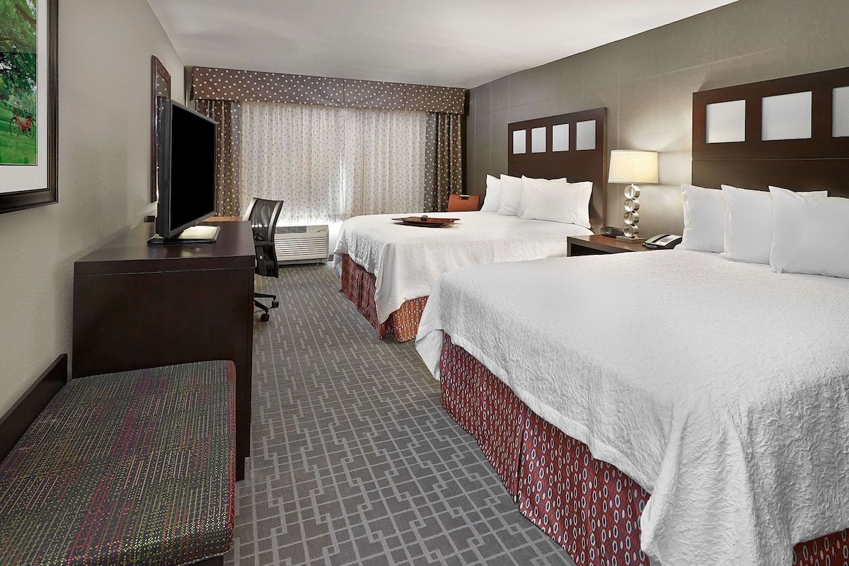 A hotel room with two beds, a desk, a chair, a TV, and a window with curtains.