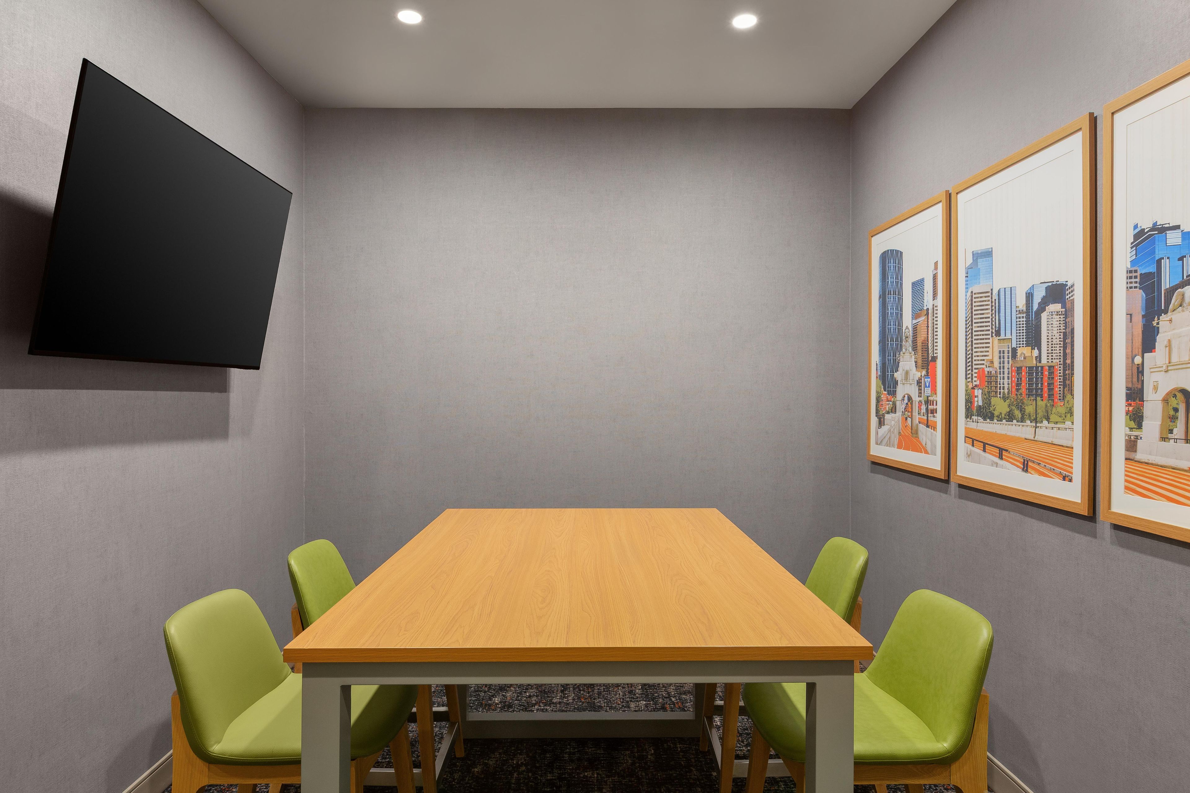 Une salle de conférence avec une table en bois, quatre chaises vertes, un téléviseur à écran plat fixé au mur et trois tableaux représentant des paysages urbains, encadrés.