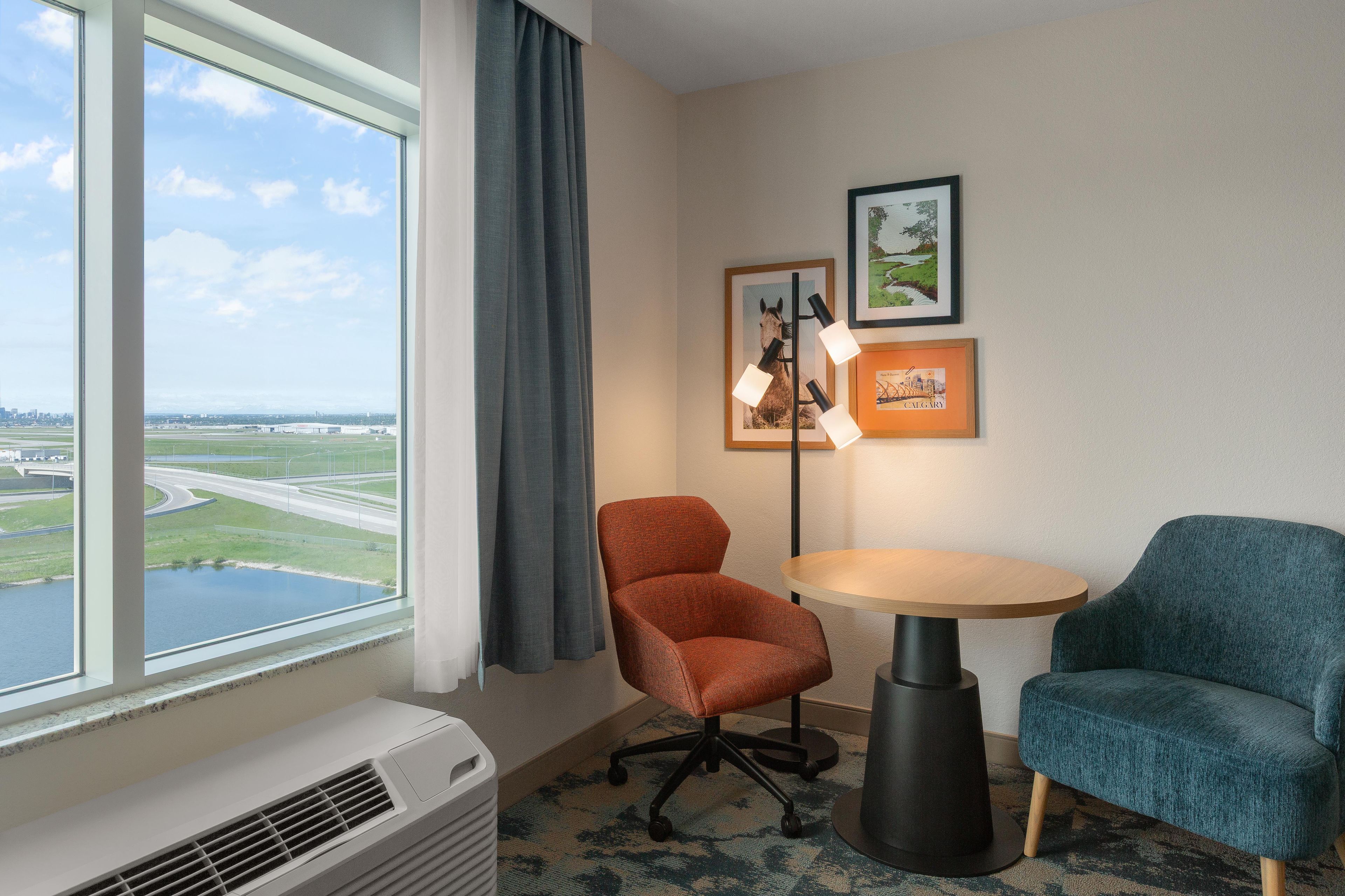 A hotel room with a large window, a desk with a lamp, two chairs, and two framed pictures on the wall.