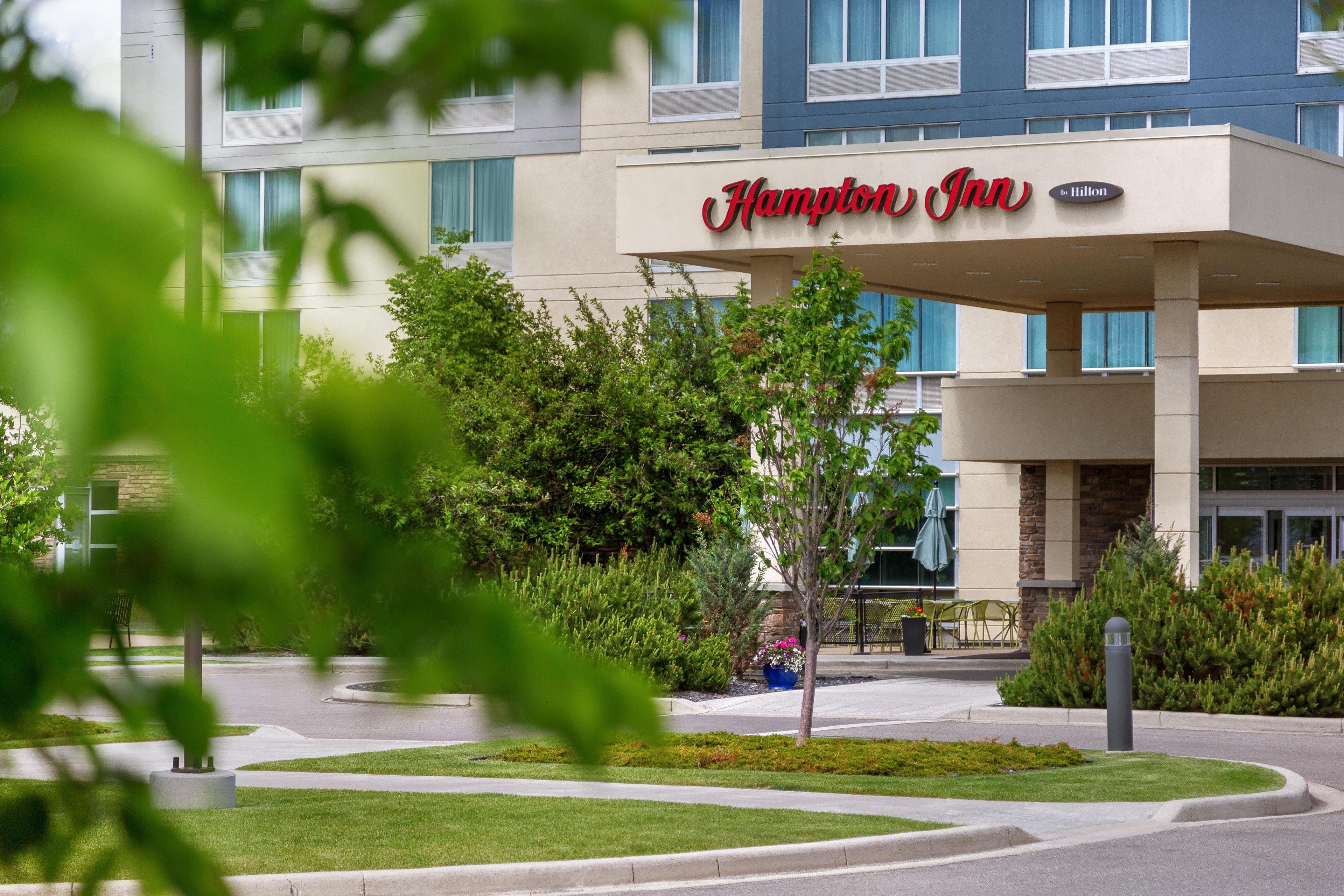 A Hampton Inn by Hilton hotel with a modern facade, a landscaped entrance, and a seating area with outdoor furniture.