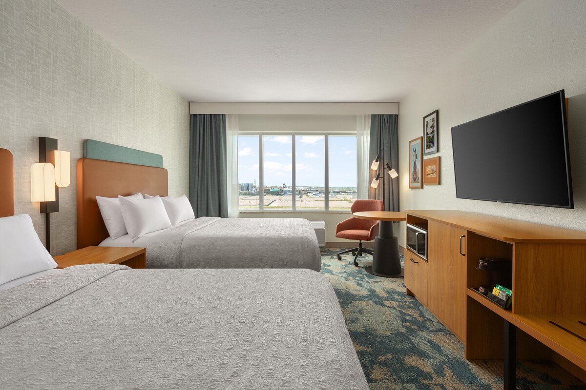 A hotel room with two beds, a desk, a television, and a view of the city.