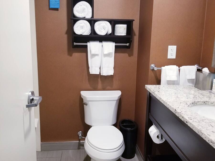 A bathroom with a white toilet, a black trash bin, a towel rack with towels, a soap dispenser, and a marble countertop with a sink.