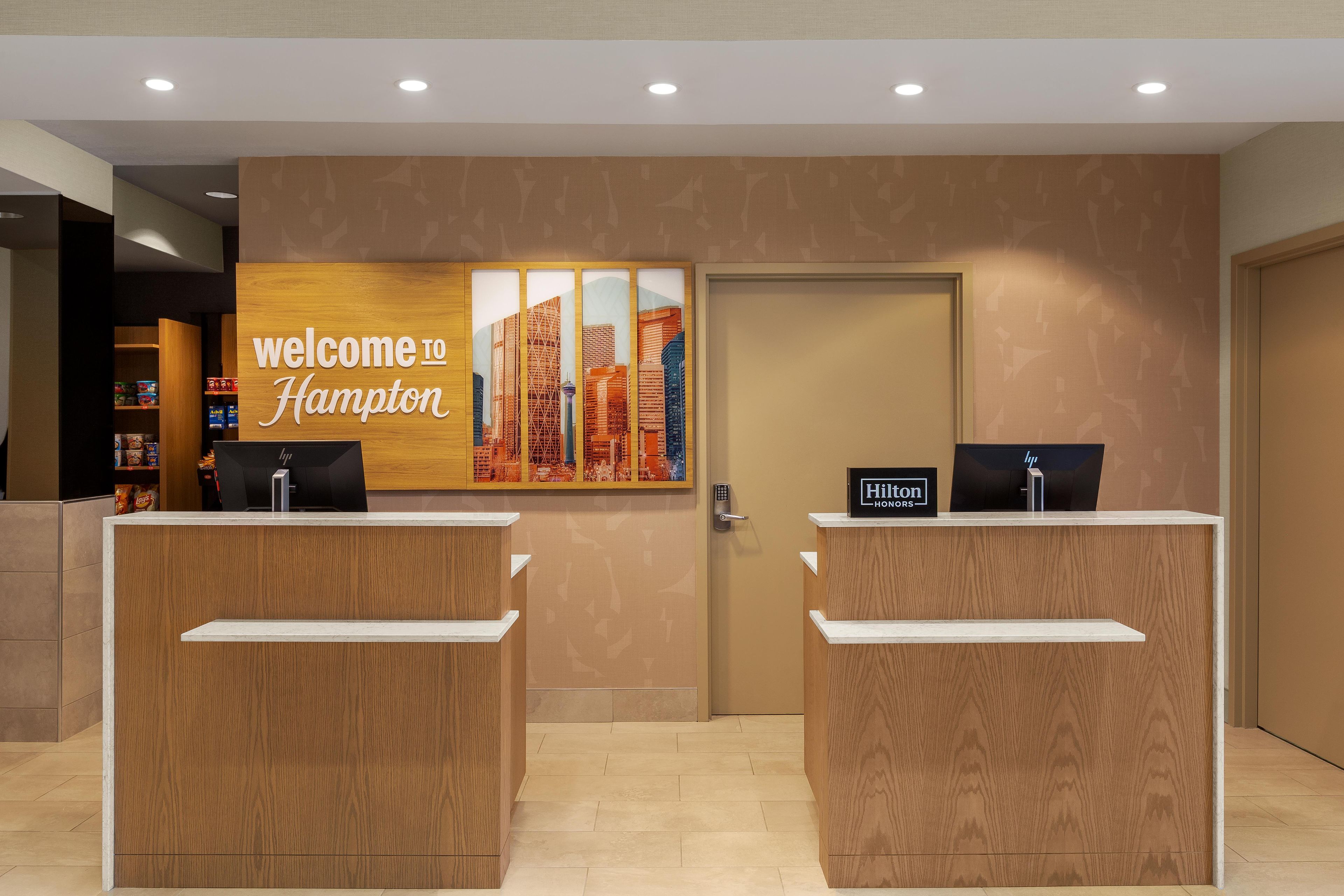 Zona de recepción de un hotel con dos mostradores, un cartel de 'Welcome to Hampton' y una obra de arte con un paisaje urbano en la pared.