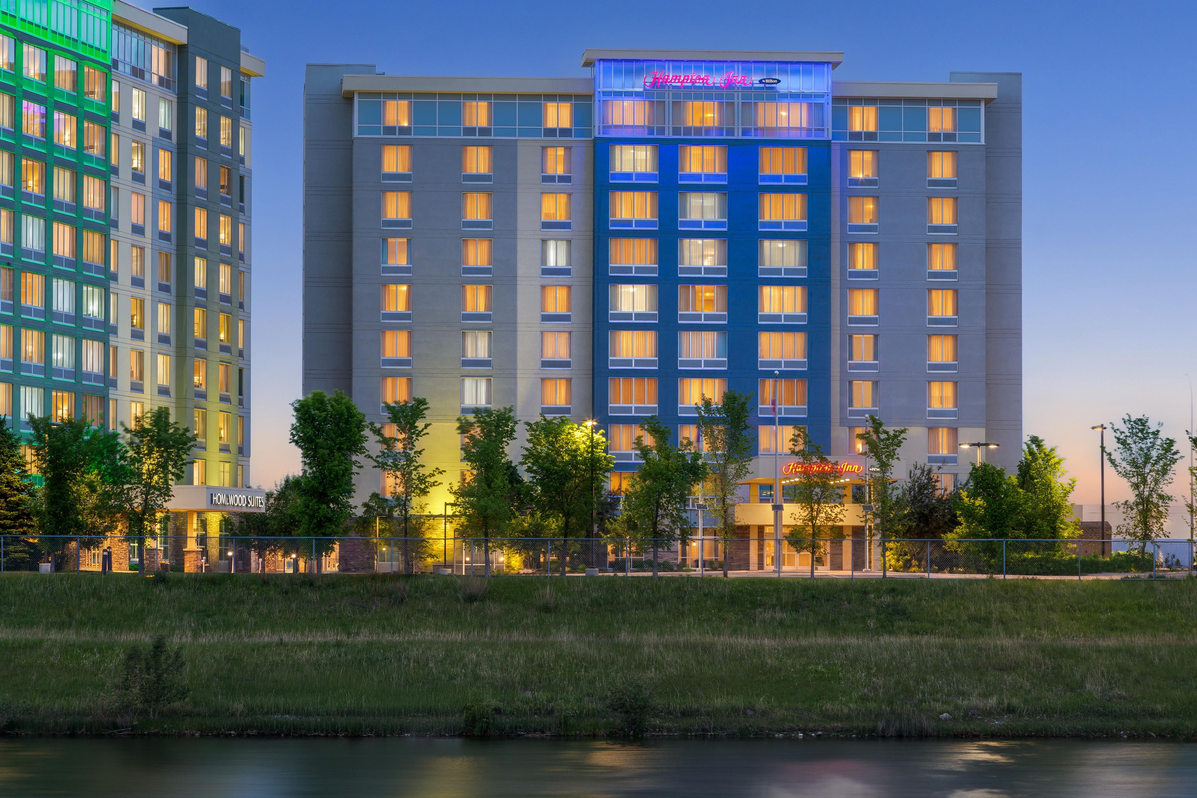 A multi-story hotel with a prominent 'Hampton Inn' sign, surrounded by greenery and a body of water.