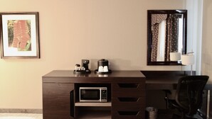 Una habitación de hotel con un escritorio de madera oscura, microondas, cafetera, una silla, un cubo de basura, un cuadro en la pared y una ventana con cortinas.