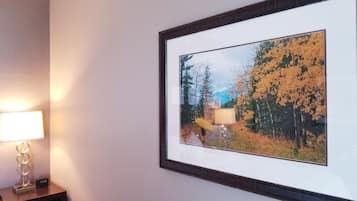 A framed picture of a forest scene with a person holding a yellow umbrella, a bedside lamp, and a digital clock on a bedside table.