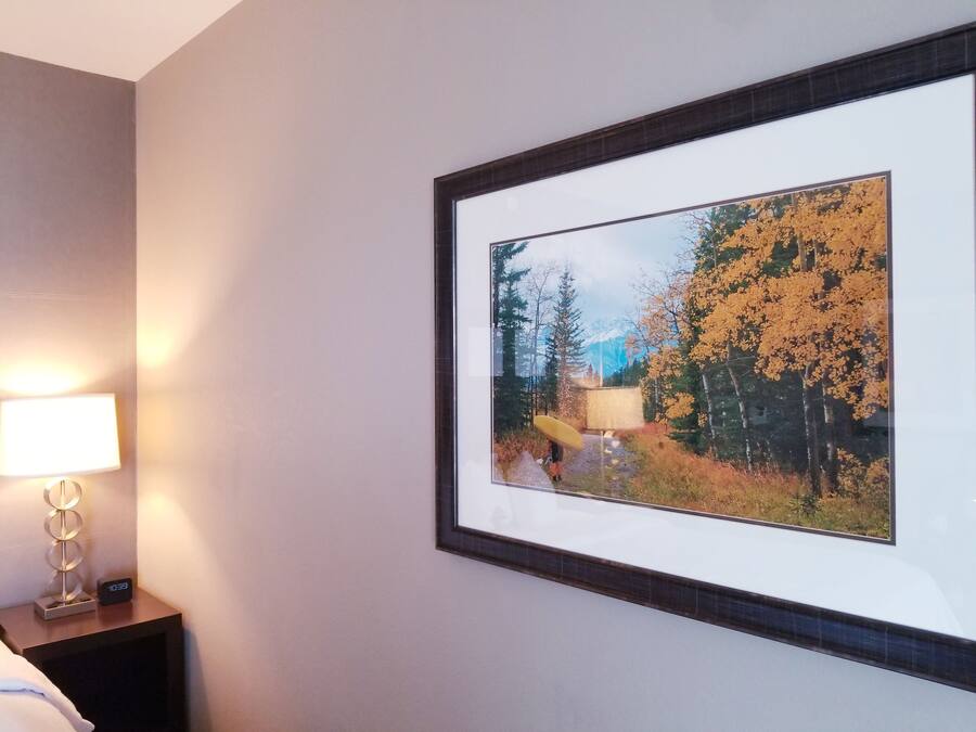 A framed picture of a forest scene with a person holding a yellow umbrella, a bedside lamp, and a digital clock on a bedside table.