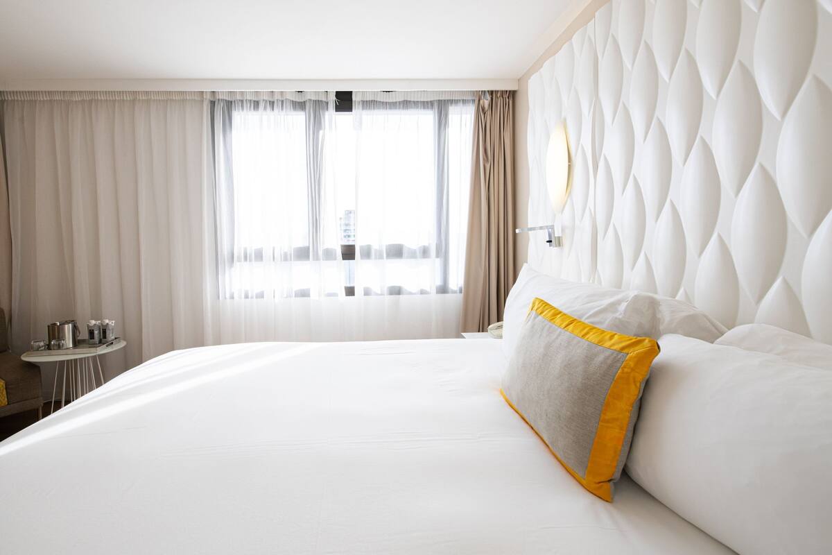A hotel room with a large bed, a small table with a lamp, and a view of the city through large windows.