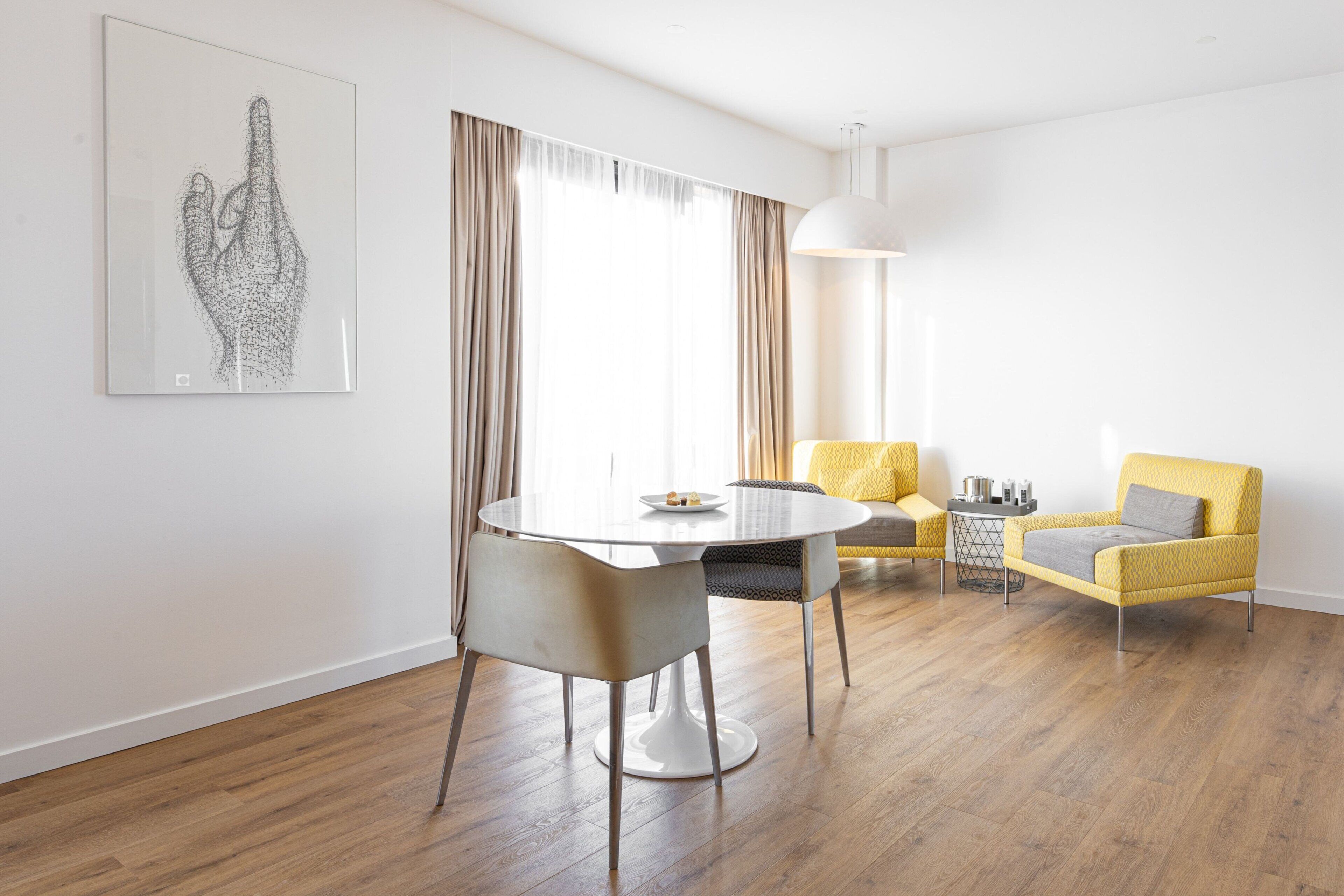 A modern living room with a round dining table, two chairs, a yellow armchair, and a large window with curtains.