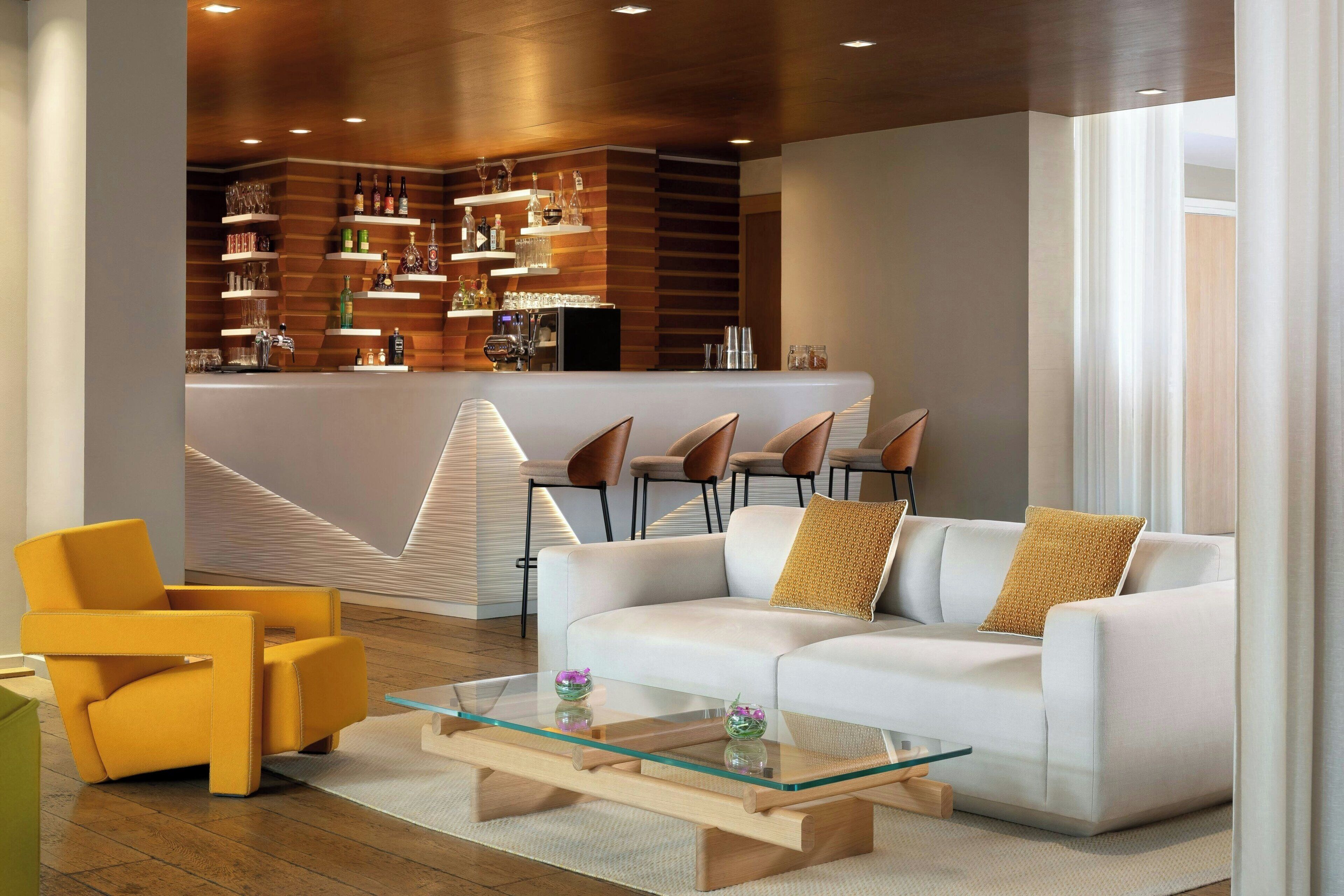 A modern living area with a white sofa, yellow armchair, and glass coffee table. A bar area with shelves of bottles and glasses is visible in the background.