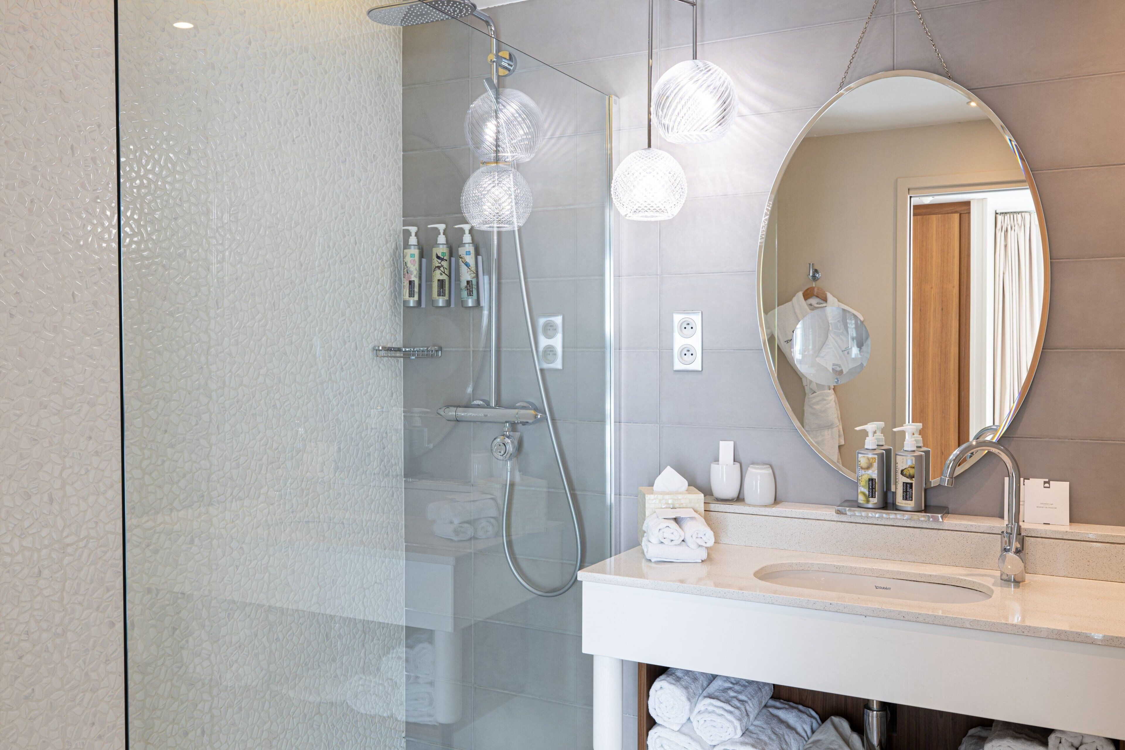 A modern bathroom with a glass shower enclosure, a round mirror, and a white sink with toiletries.