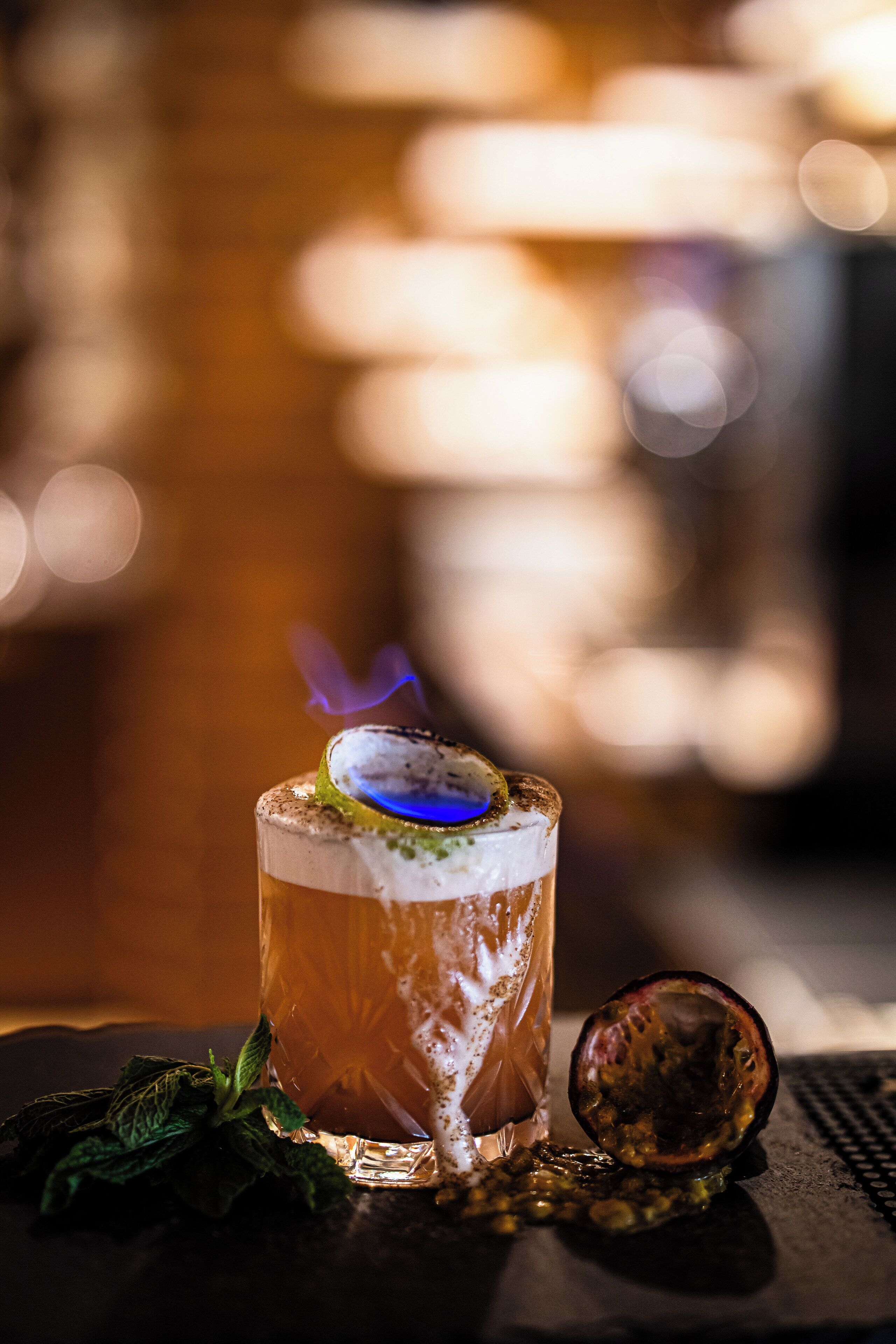 Cocktail-drinkki, jossa on limenviipale ja sininen liekki koristeena, ja se on koristeltu mintunlehdillä, ja se on tarjoiltu baaritiskillä.