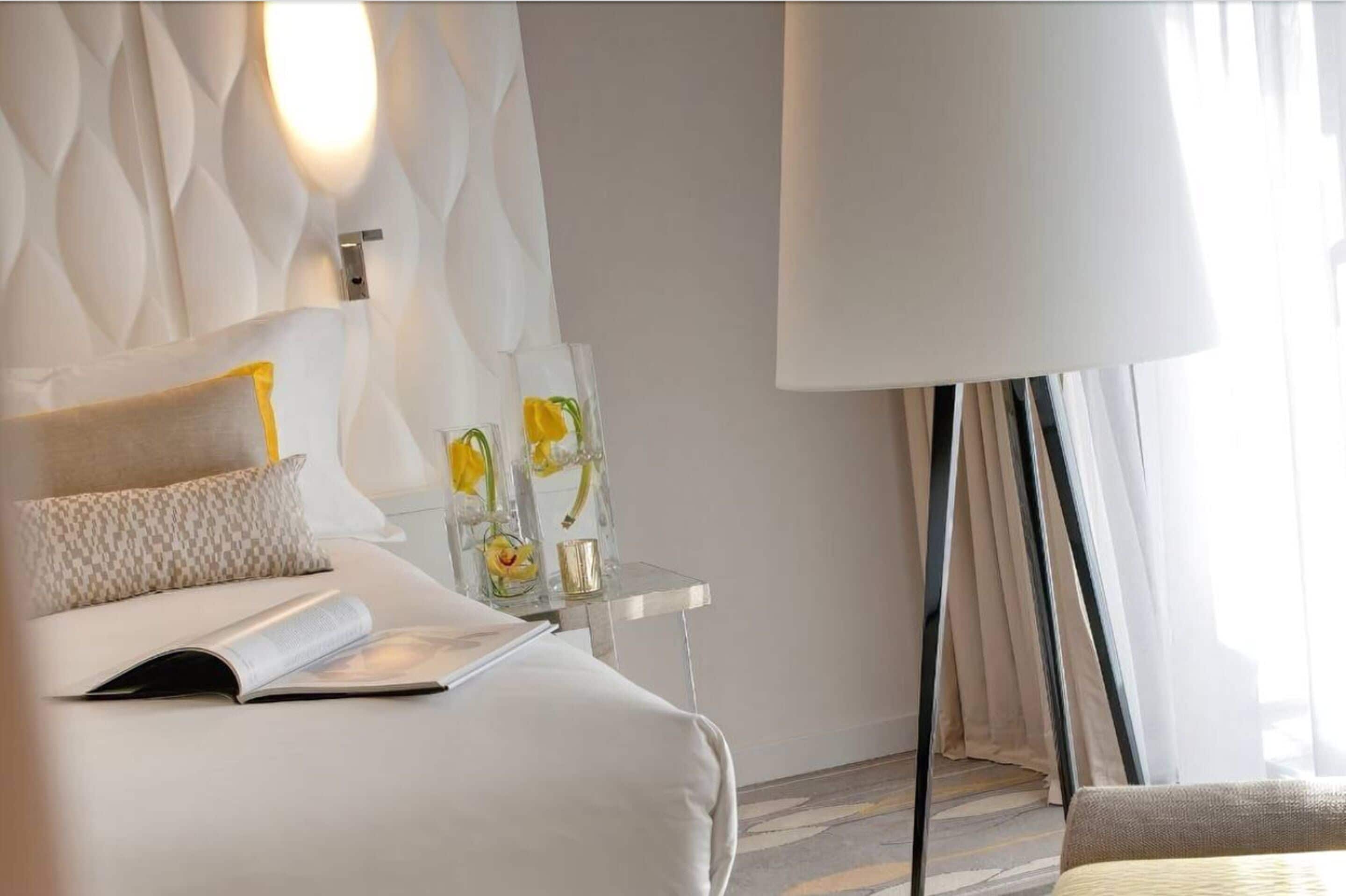 A hotel room with a bed, a bedside table with decorative items, a lamp, and a view of the outdoors.