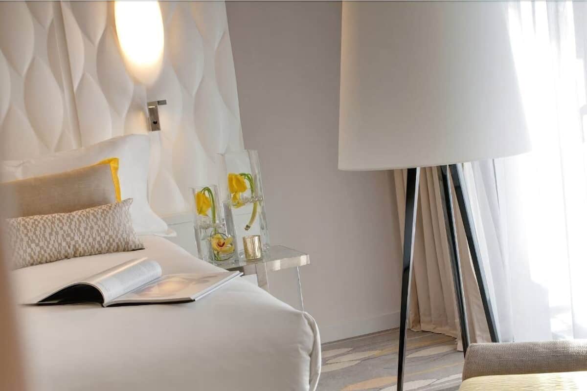A hotel room with a bed, a bedside table with decorative items, a lamp, and a view of the outdoors.
