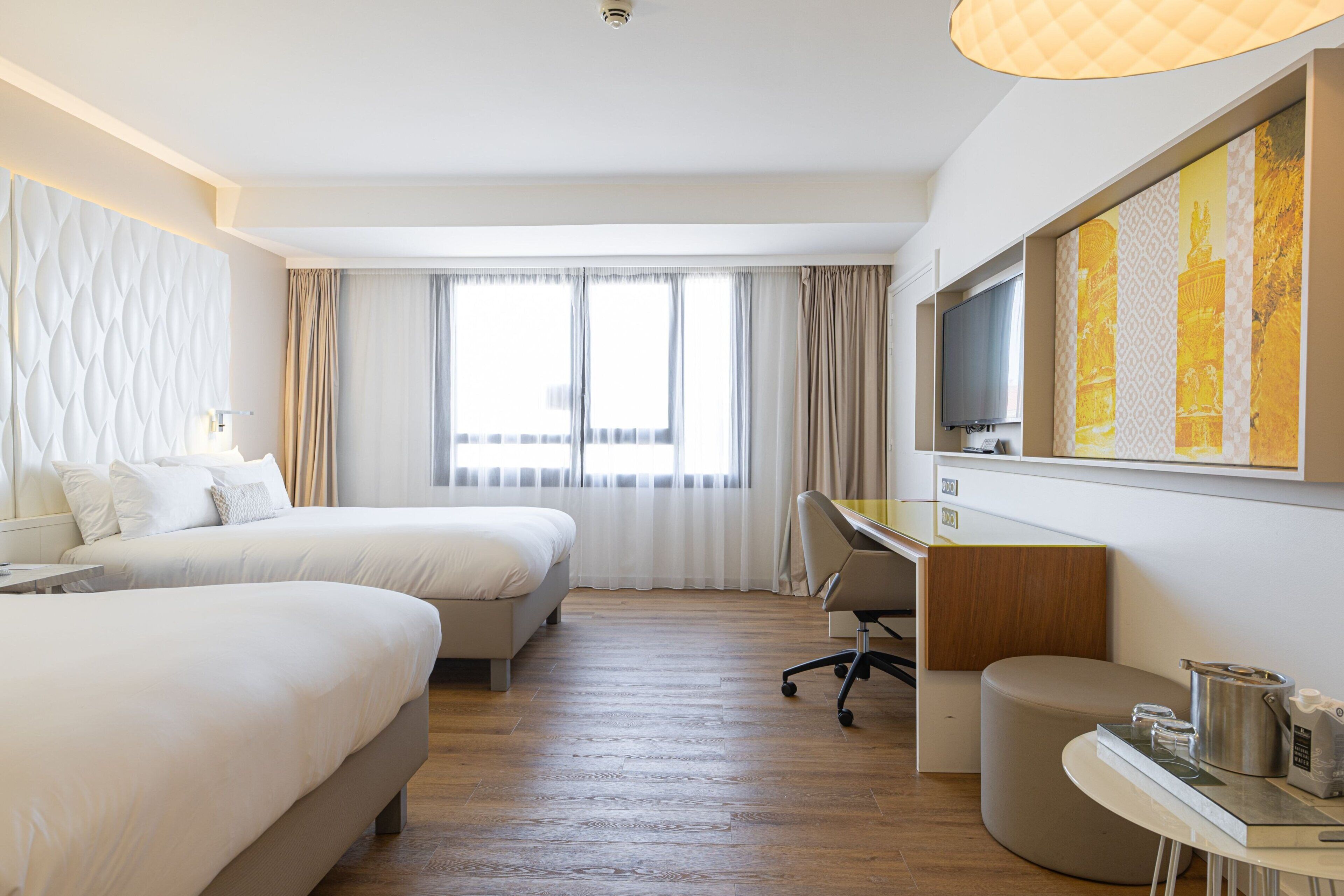 A hotel room with two beds, a desk, a chair, a television, and a large window with curtains.