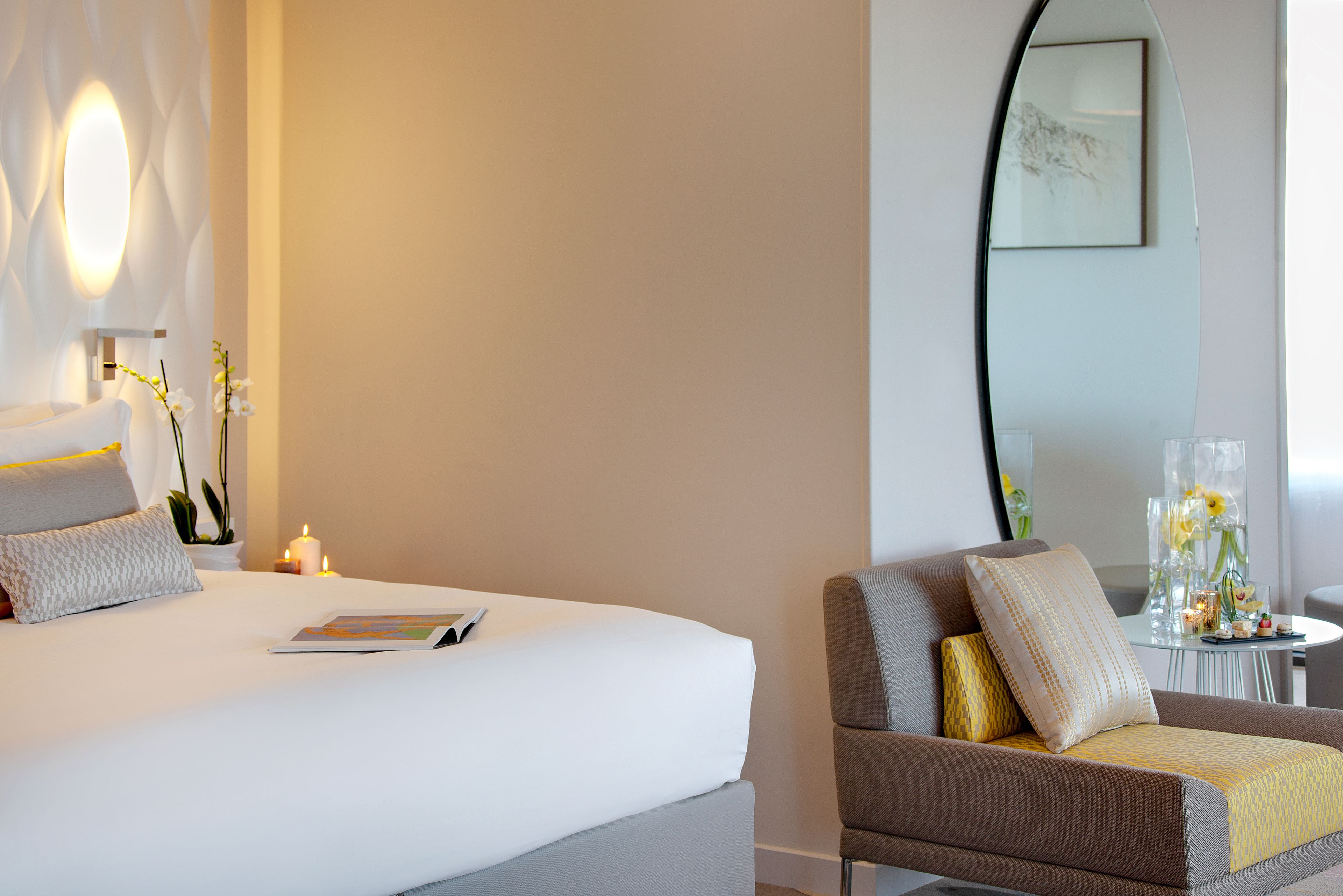 A hotel room with a bed, a bedside table with a candle, a chair with cushions, and a small table with decorative items.