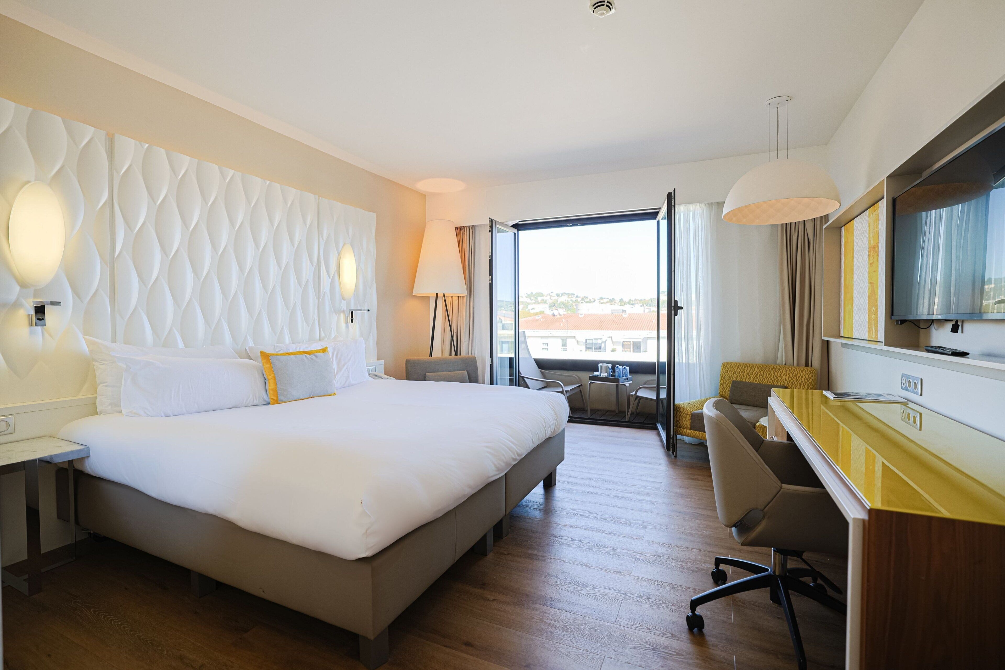 Habitación de hotel con una cama grande, un escritorio, una silla, un televisor y vista a la ciudad.