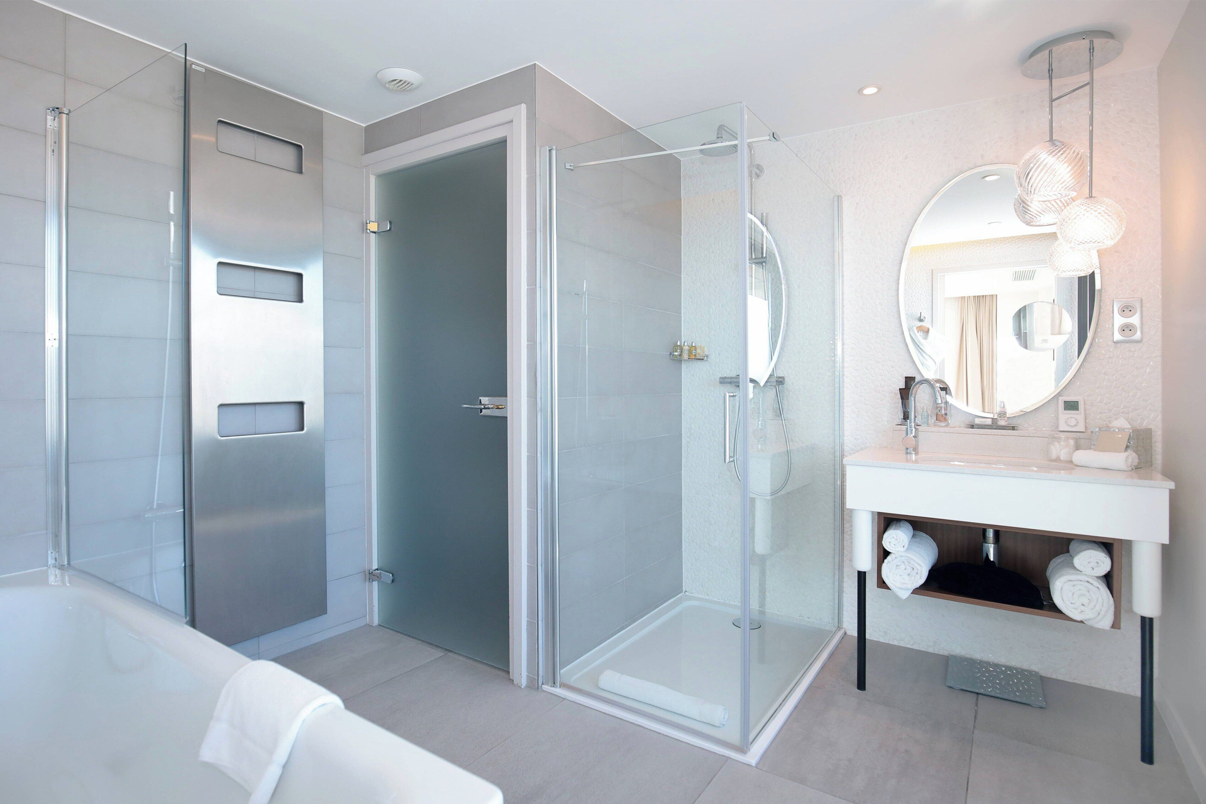 A modern bathroom with a glass-enclosed shower, a white bathtub, and a vanity with a round mirror.