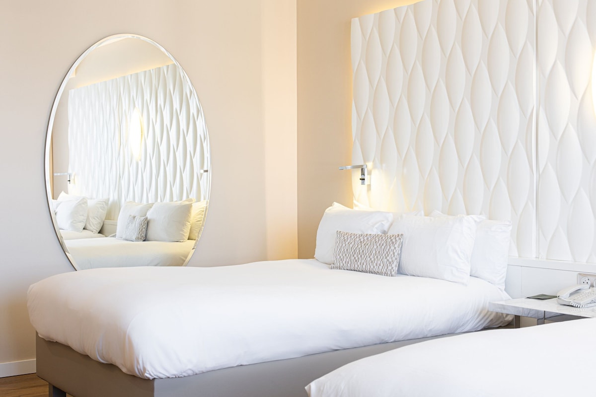 Une chambre d’hôtel moderne avec un grand miroir rond, une tête de lit capitonnée, un lit bien fait avec du linge de lit blanc, et une table de chevet avec une lampe et un téléphone.
