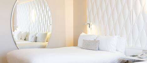 A modern hotel room with a large round mirror, a tufted headboard, a neatly made bed with white linens, and a bedside table with a lamp and a phone.