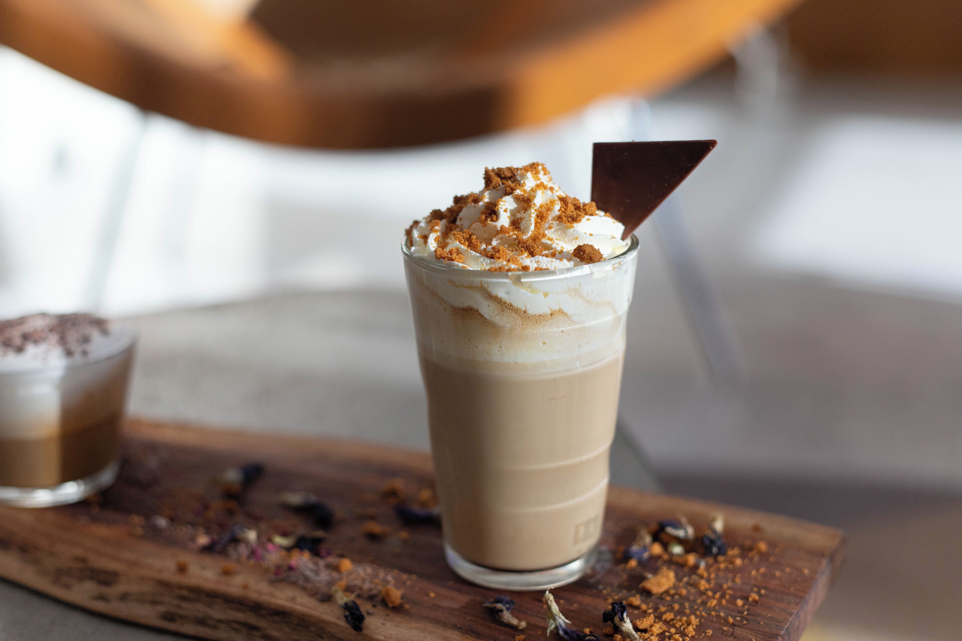 Un verre contenant une boisson à plusieurs couches, surmontée de crème fouettée et d’un morceau de chocolat, posé sur une planche en bois parsemée de miettes.