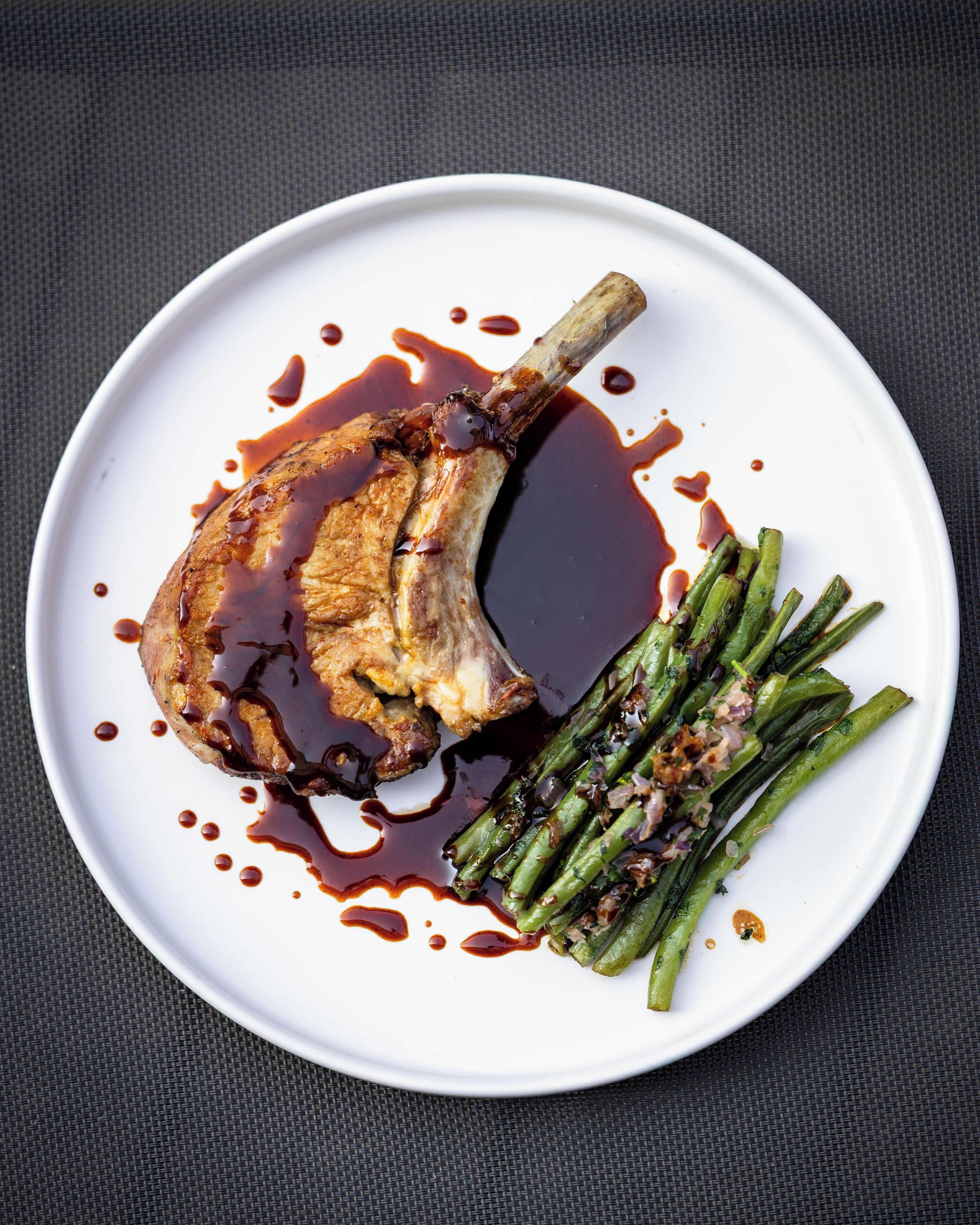 Une assiette blanche contenant une côte d’agneau grillée, des asperges et une sauce foncée.