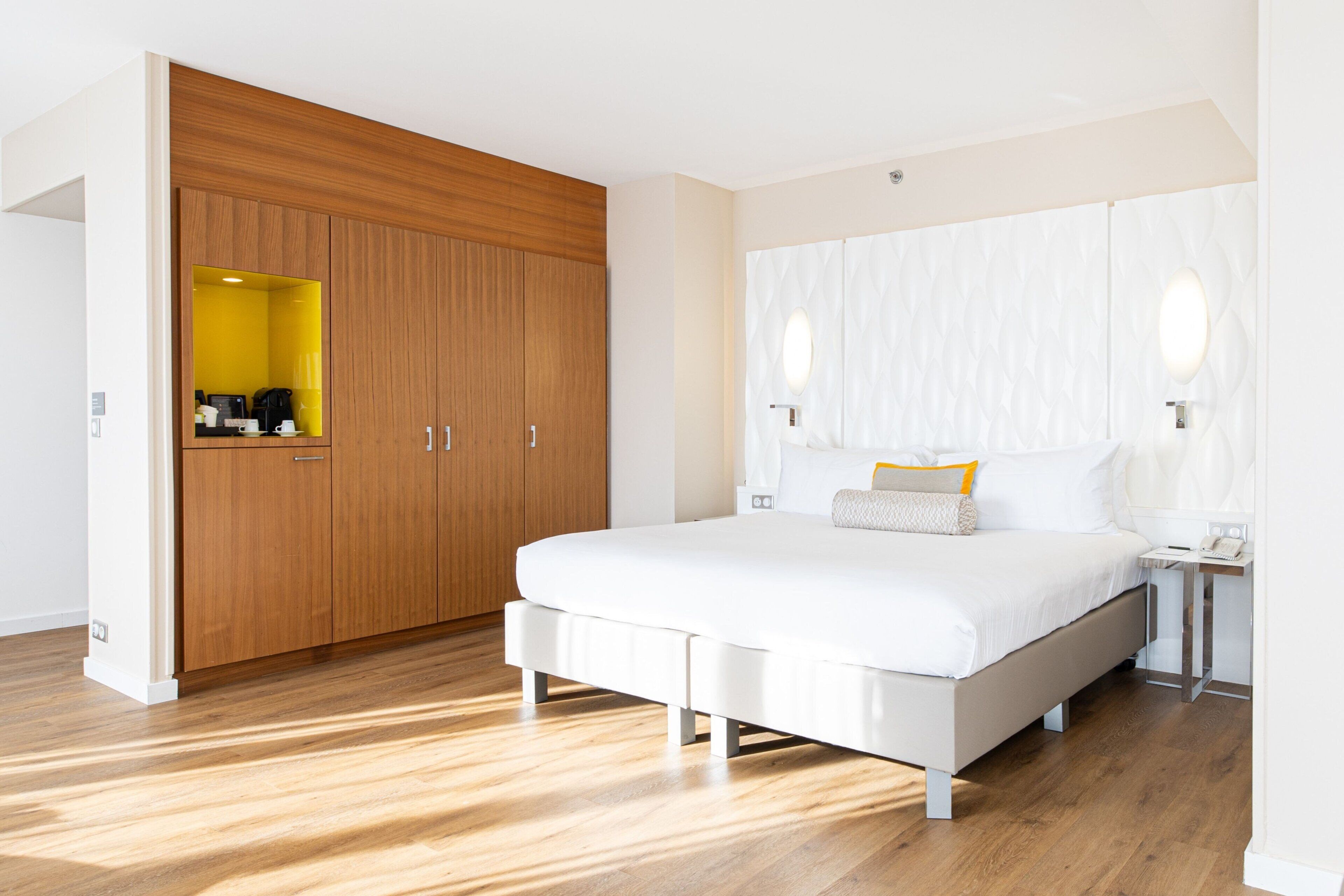 Une chambre à coucher moderne avec un grand lit, une armoire en bois et une petite salle de bain dotée d’un miroir et d’un luminaire.