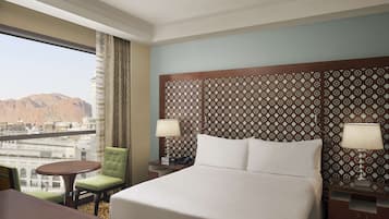 A hotel room with a large bed, a dining table, a green chair, and a view of mountains through the window.